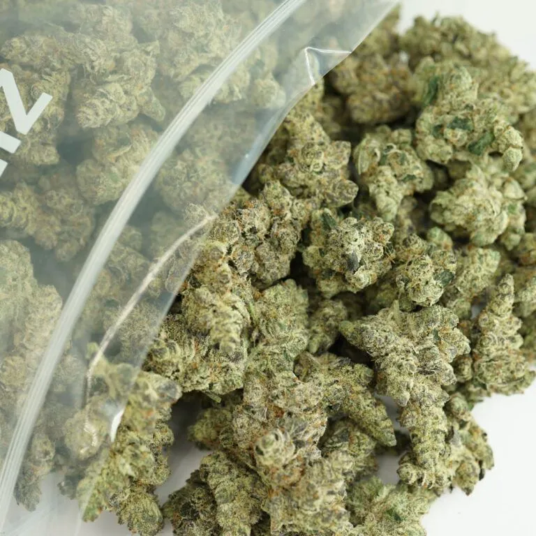 Large clear bag filled with dense cannabis buds, showcasing frosty green and orange tones with visible trichomes on a white background.