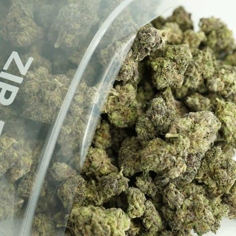 close-up DOMAMINE BULK image of cannabis buds spilling out of a clear ZIP-ZAG plastic bag onto a white surface, showing dense green nugs with visible trichomes and orange hairs.