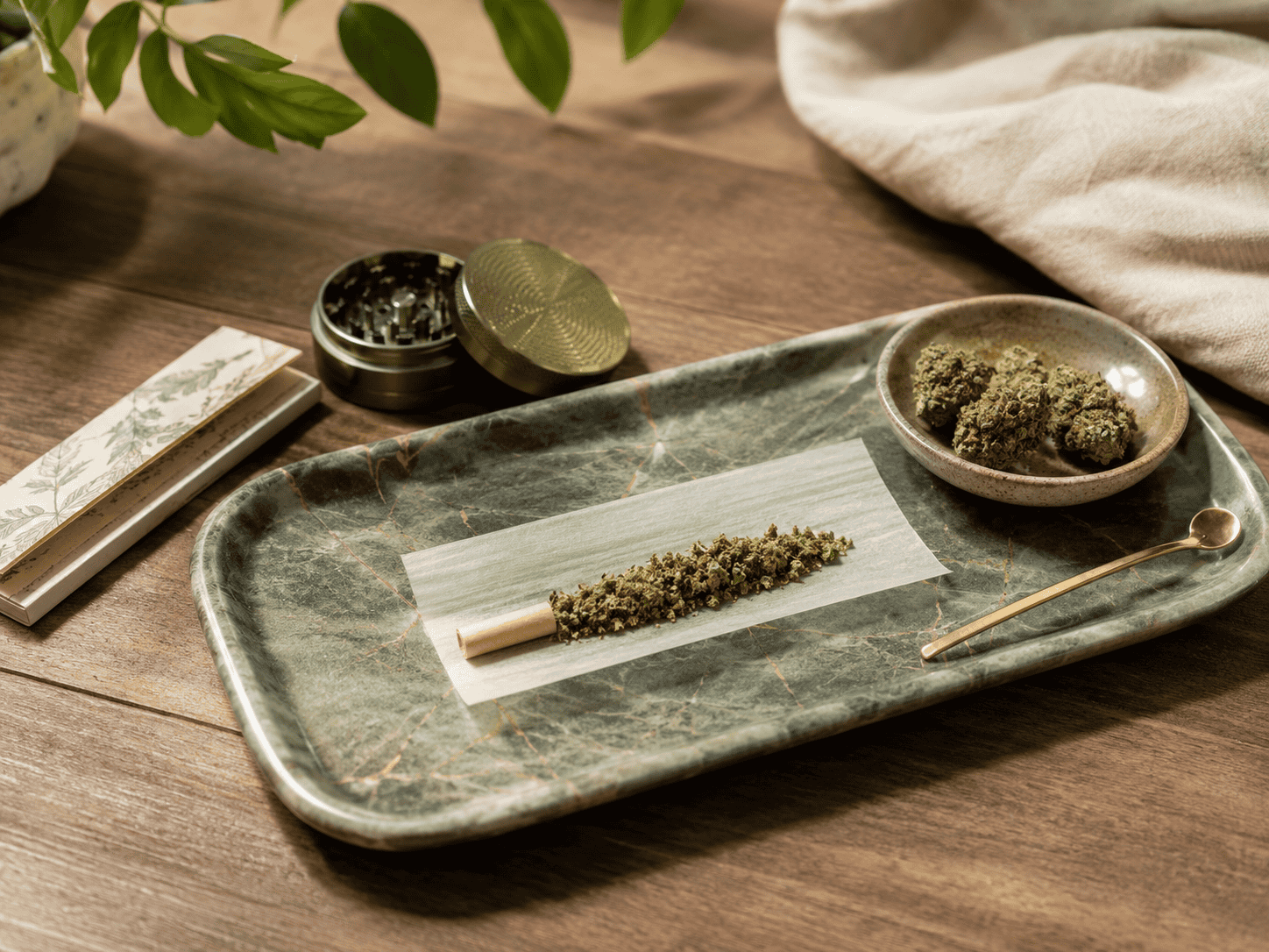 Cannabis rolling tray with a rolling paper, ground cannabis flower, grinder, and packing tool arranged on a wooden tabletop.