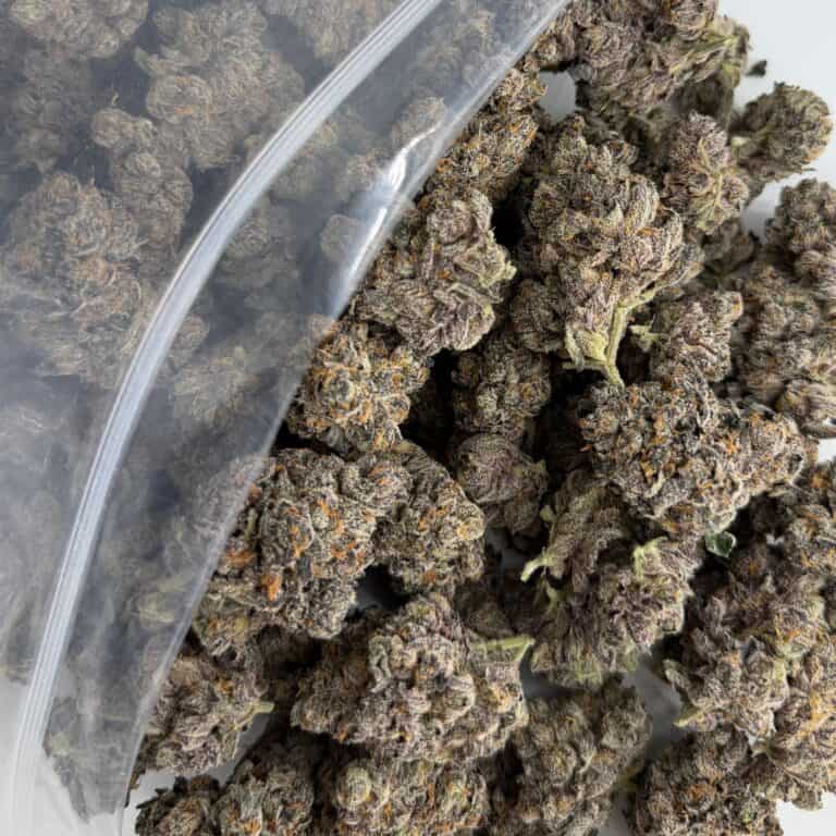 close-up image of cannabis buds spilling out of a clear ZIP-ZAG plastic bag onto a white surface, showing dense green nugs with visible trichomes and orange hairs.