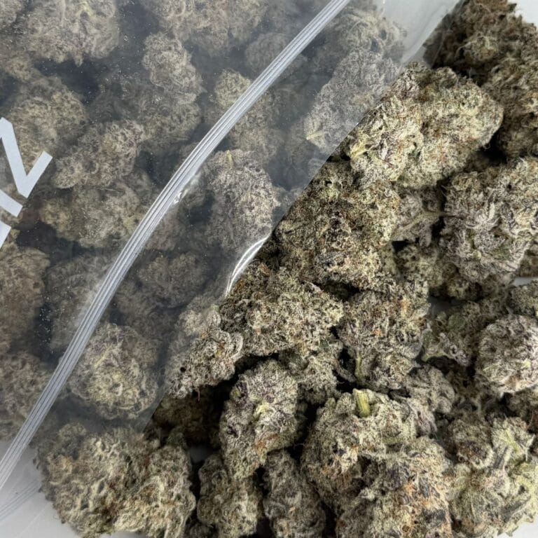 close-up image of cannabis buds spilling out of a clear ZIP-ZAG plastic bag onto a white surface, showing dense green nugs with visible trichomes and orange hairs.
