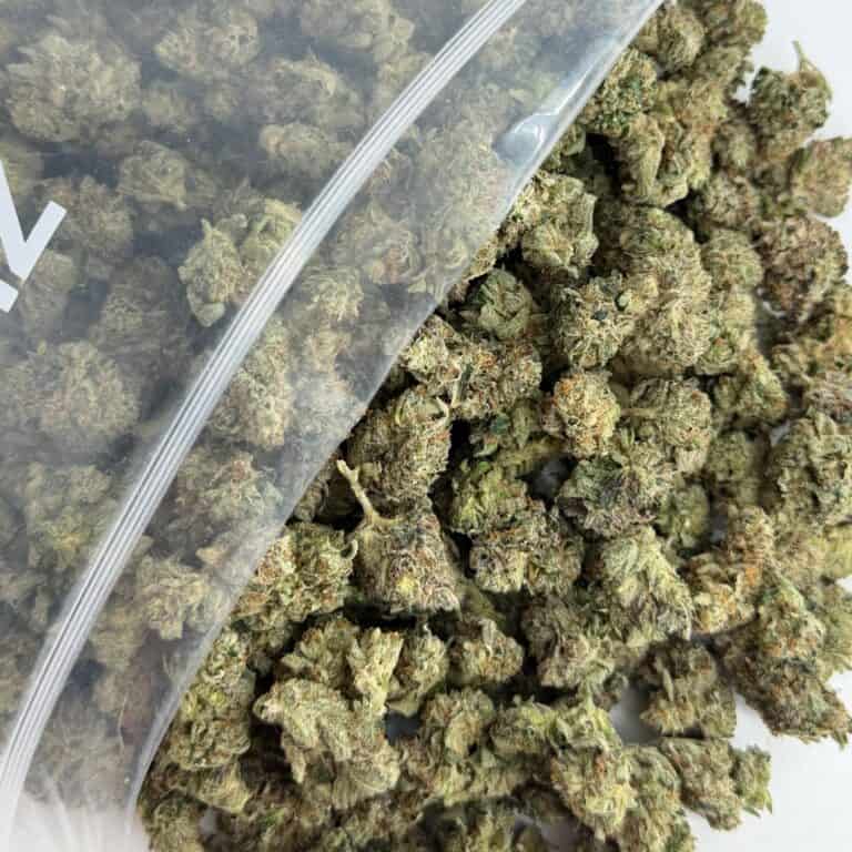 close-up image of cannabis buds spilling out of a clear ZIP-ZAG plastic bag onto a white surface, showing dense green nugs with visible trichomes and orange hairs.