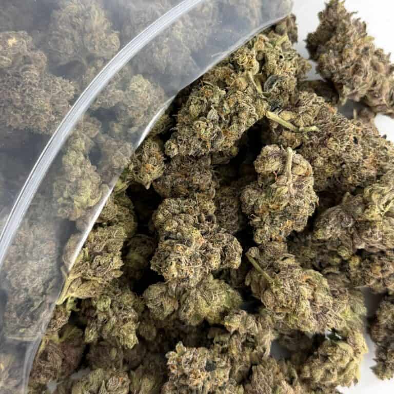 close-up image of cannabis buds spilling out of a clear ZIP-ZAG plastic bag onto a white surface, showing dense green nugs with visible trichomes and orange hairs.