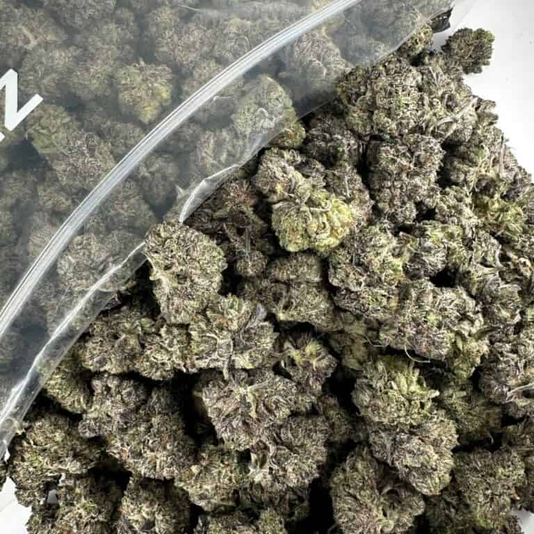 close-up image of cannabis buds spilling out of a clear ZIP-ZAG plastic bag onto a white surface, showing dense green nugs with visible trichomes and orange hairs.