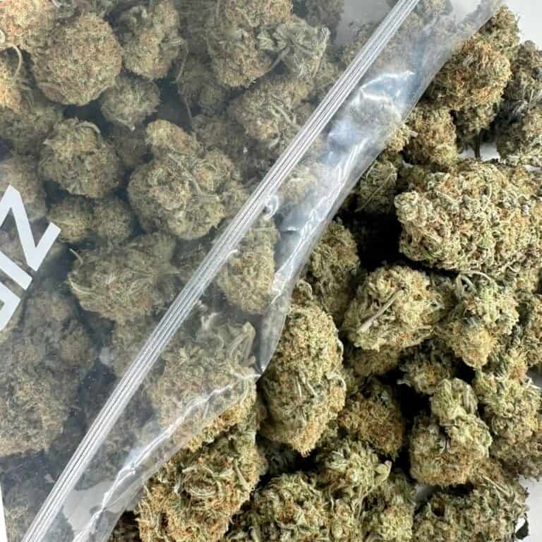 close-up image of cannabis buds spilling out of a clear ZIP-ZAG plastic bag onto a white surface, showing dense green nugs with visible trichomes and orange hairs.